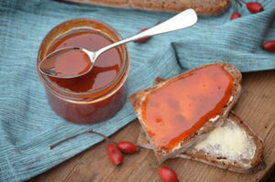 Brot mit Hagebutten-Marmelade und Marmeladen-Glas mit Löffel auf blauer Tischdecke.